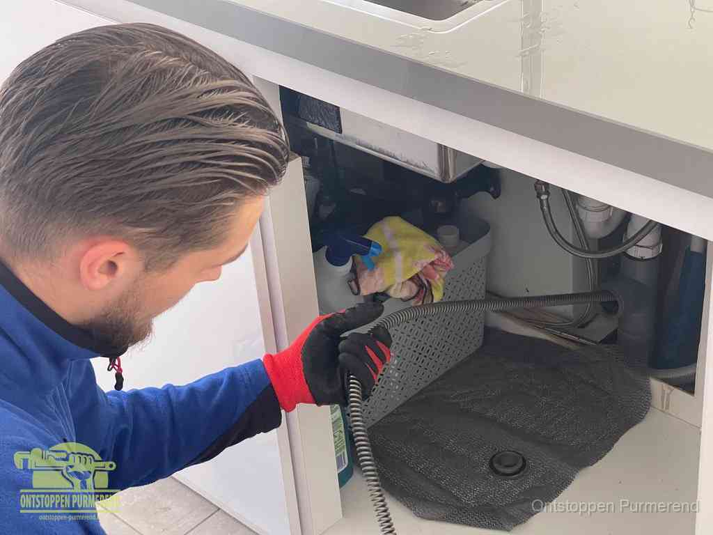 Een monteur voert een flexibele ontstoppingsveer in onder het keukenkastje, langs de sifon, om de gootsteen te ontstoppen in Purmerend