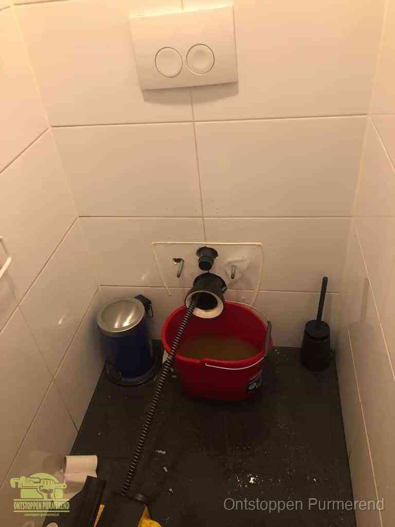 Een ontstopte wand-wc met verwijderde toiletpot waar een ontstoppingsveer in de wanduitgang verdwijnt, het achterhalen waar de verstopping zit in Purmerend