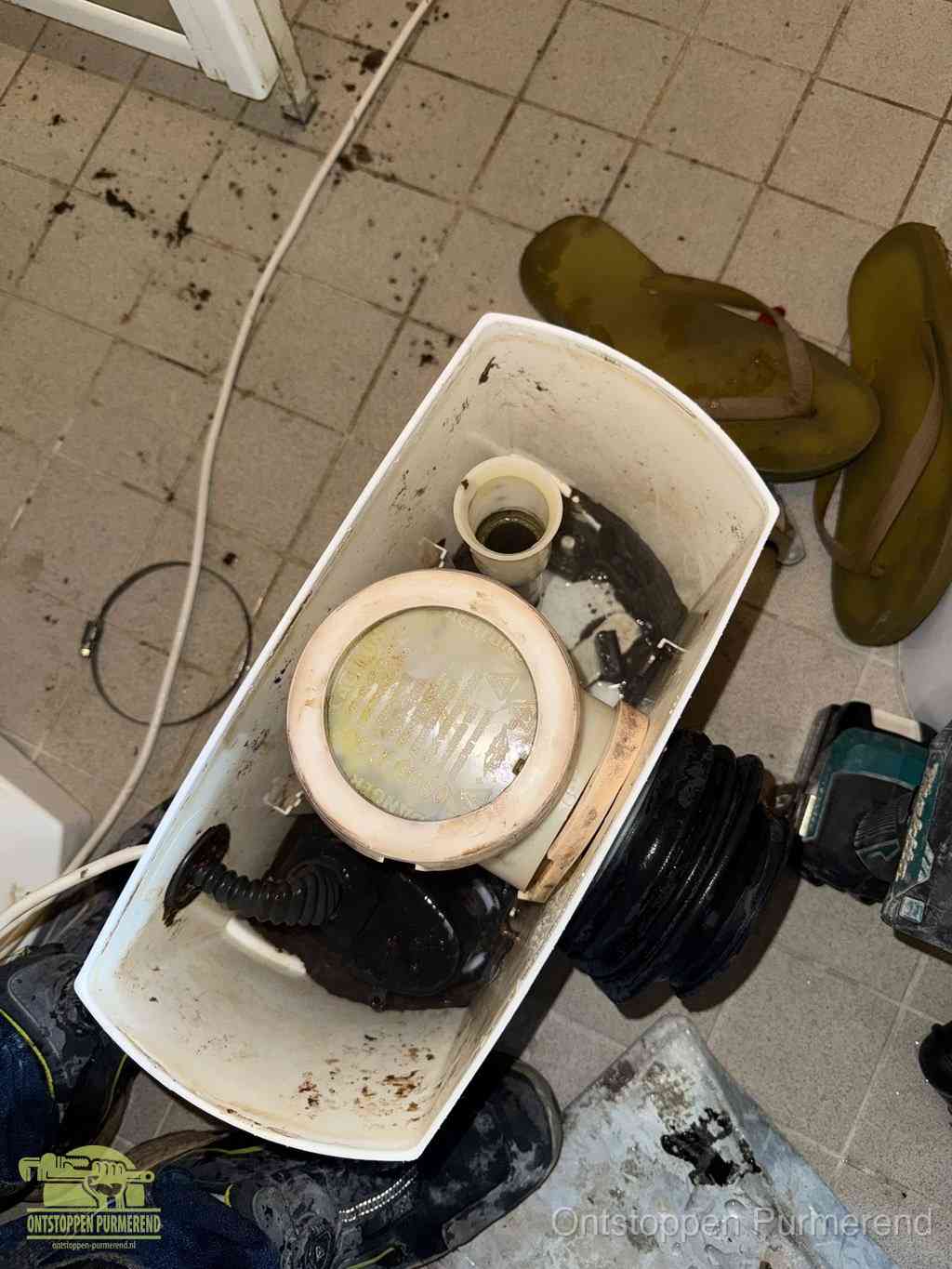 Een gedemonteerde toiletpot op de vloer met ontstoppingsveer en gereedschap ernaast, het verwijderen van een vastzittend voorwerp uit de wc in Purmerend
