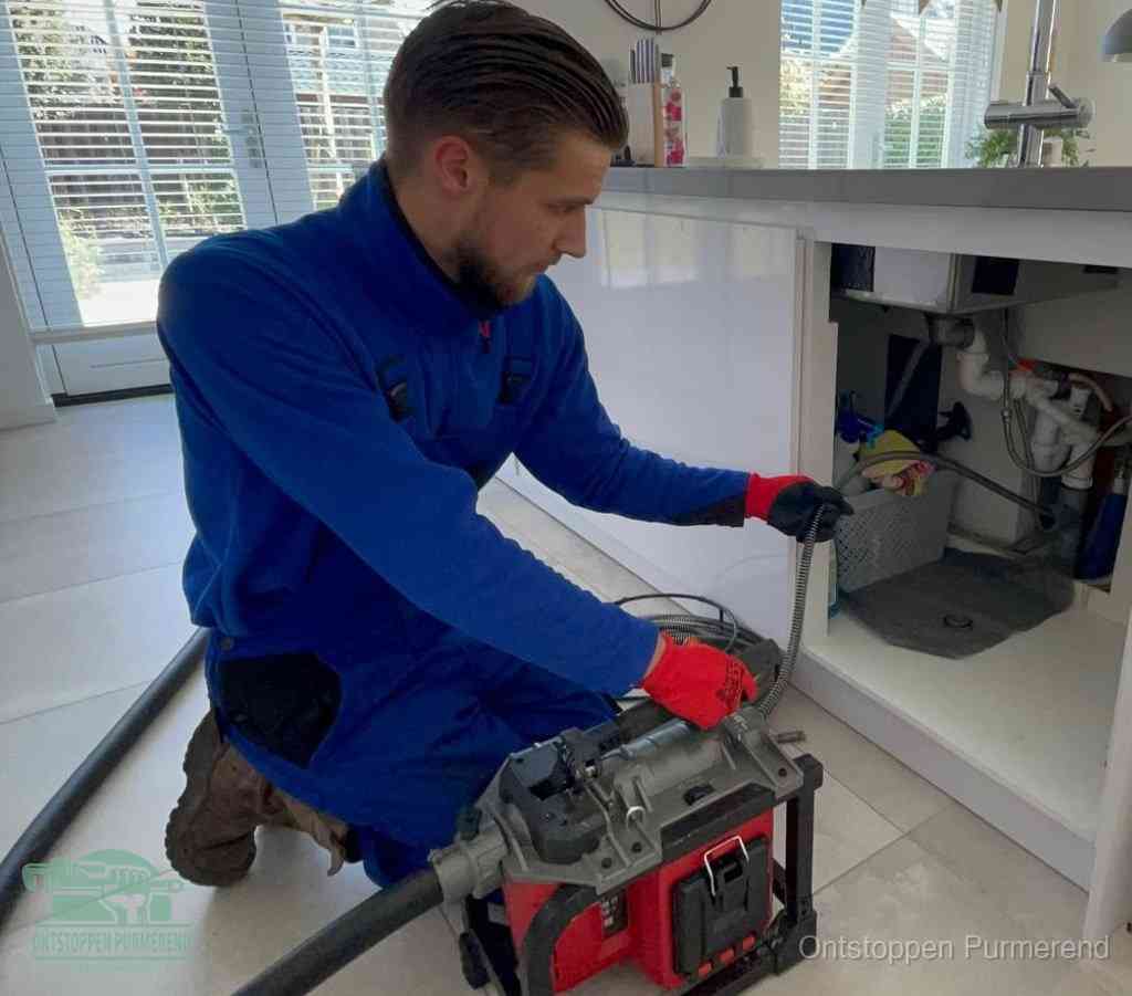 Een monteur met rode handschoenen knielt in een keuken en voert een elektrische ontstoppingsveer in tijdens het professioneel afvoer ontstoppen in Purmerend
